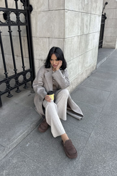 a lady in a grey patchwork top, a grey long coat and a pair of beige pants